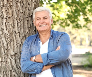 Older man smiling with arms crossed while leaning against tree